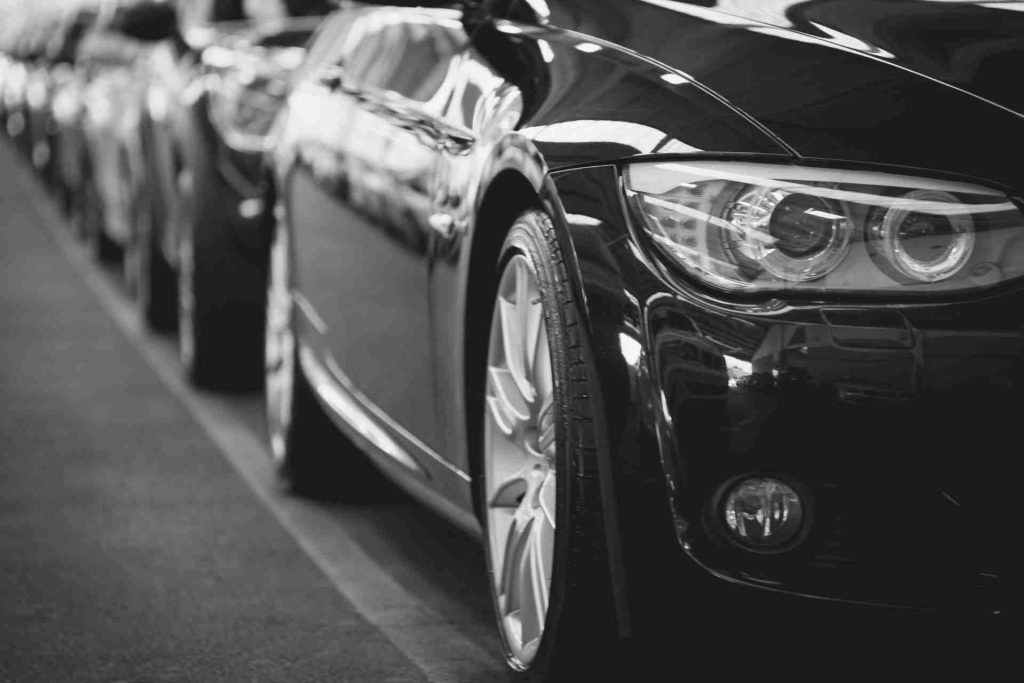 Black and white photograph depicting a row of cars parked in a lot, showcasing their shapes and shadows.