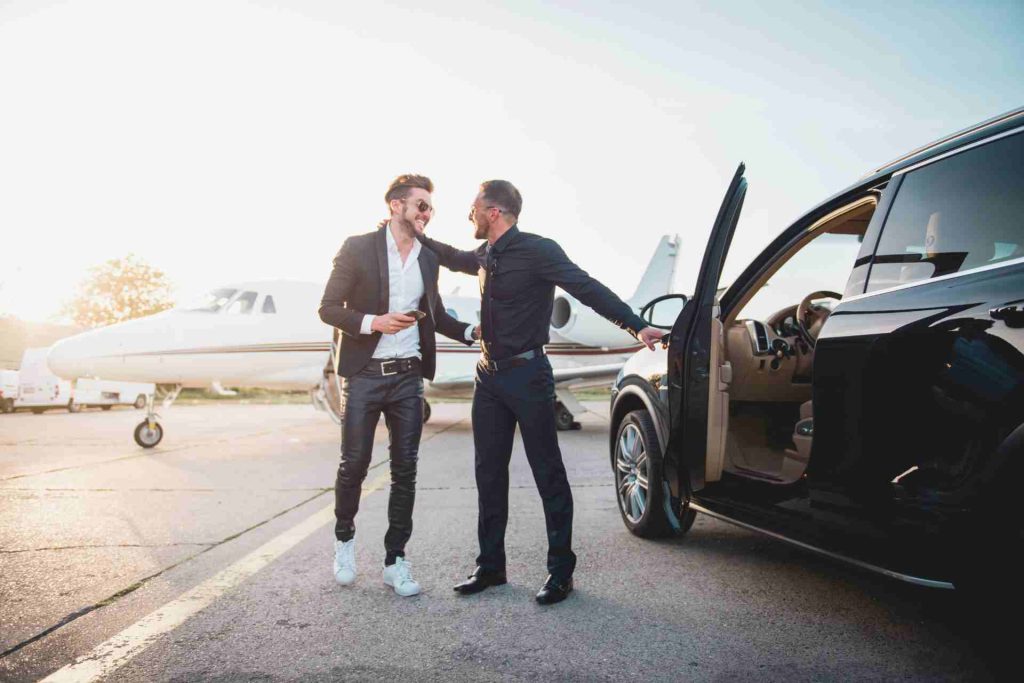 Two men stand beside a car, with a private jet visible in the background, suggesting luxury and travel.