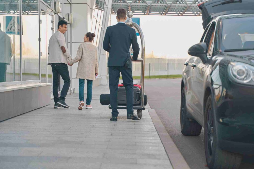 A man and woman beside a parked car, together with a driver helping with their luggage.