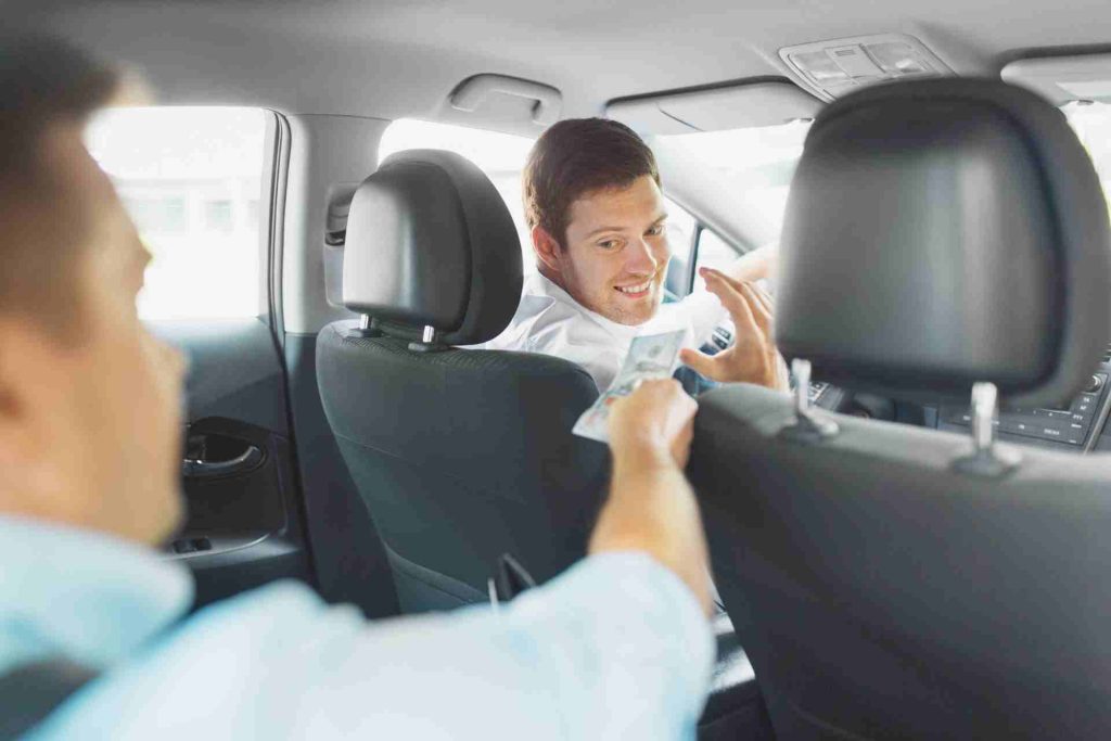 A man seated in the back seat of a car giving a tip to the driver.